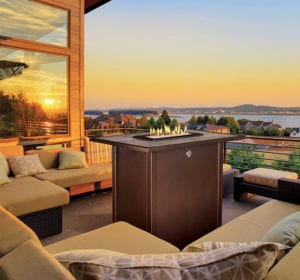 A tall, rectangular outdoor gas fireplace sits ablaze in the middle of a large deck with light brown patio furniture.