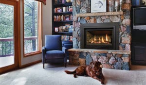 A gas fireplace insert is lit inside a mantel made of large, multi-colored stones. A brown dog relaxes in front of the fire.