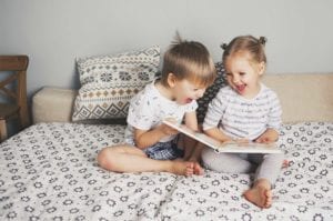 A young boy and girl sit on bed laughing and reading a book