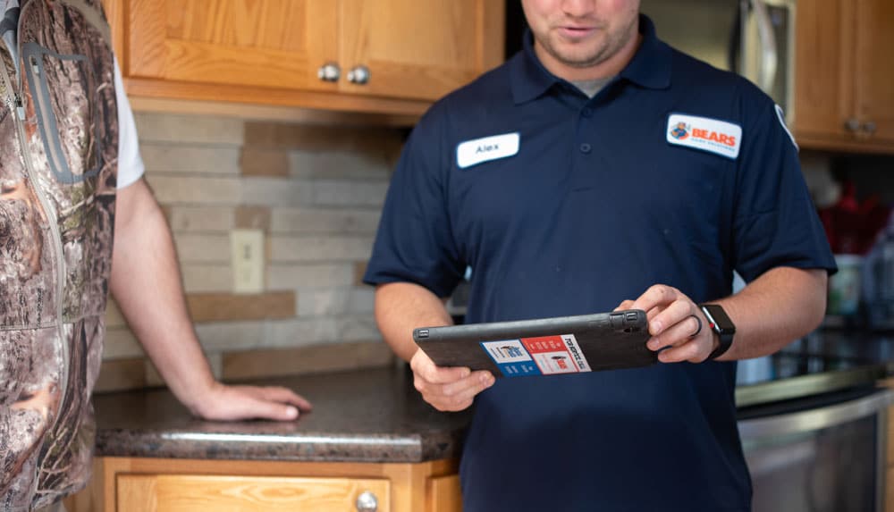 A male heating and cooling technician holding a tablet in his hands interacts with a homeowner in a kitchen