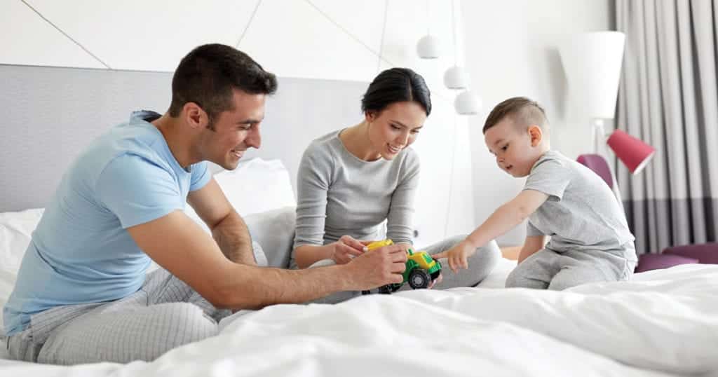 A father, mother, and son sit on a large bed and look at a toy truck together