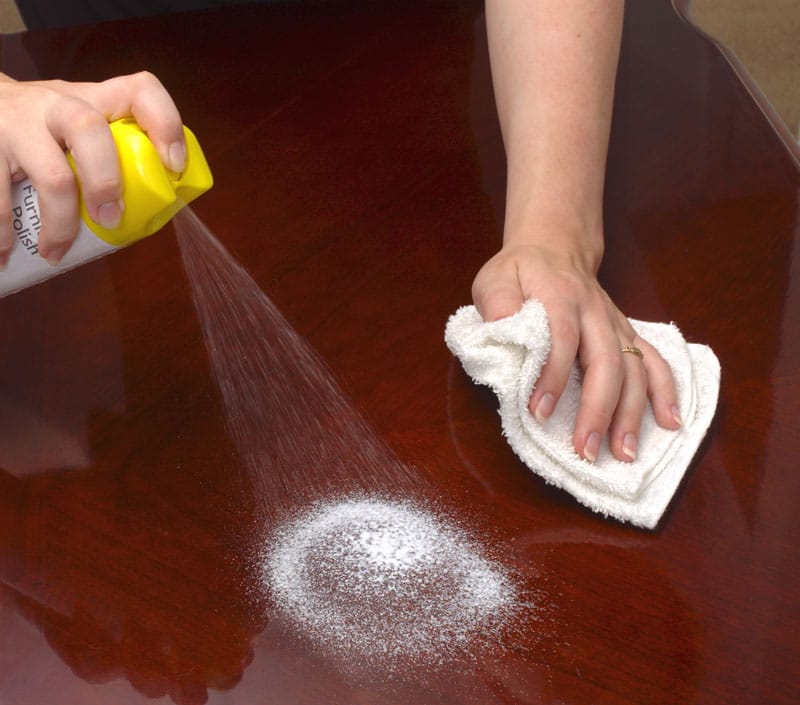 A closeup of a person’s hands spraying furniture polish on a wooden table