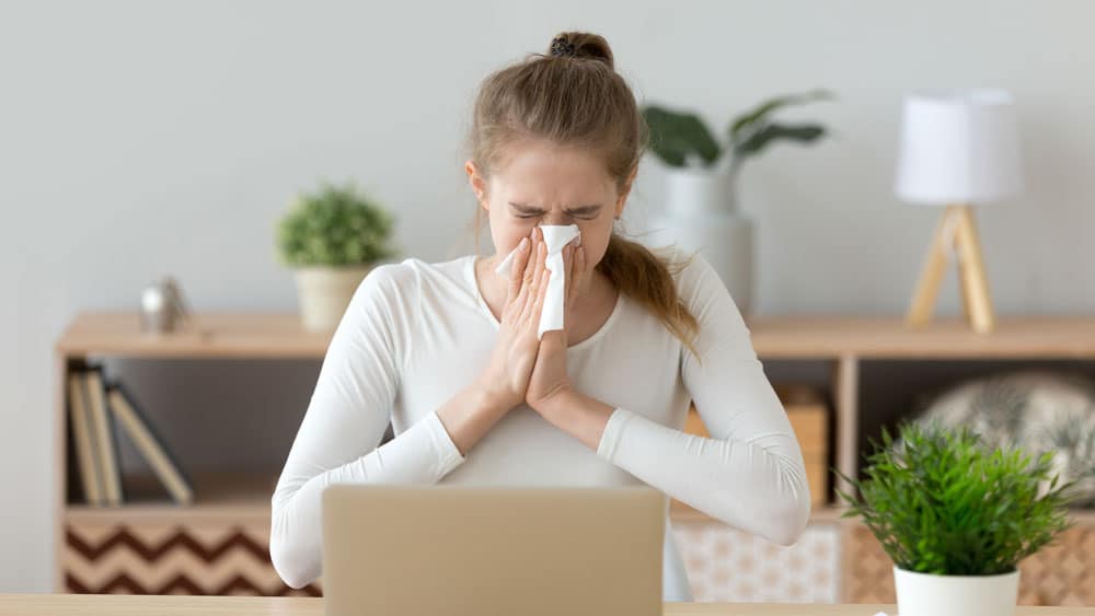 Woman sneezes into tissue while working on laptop in her home