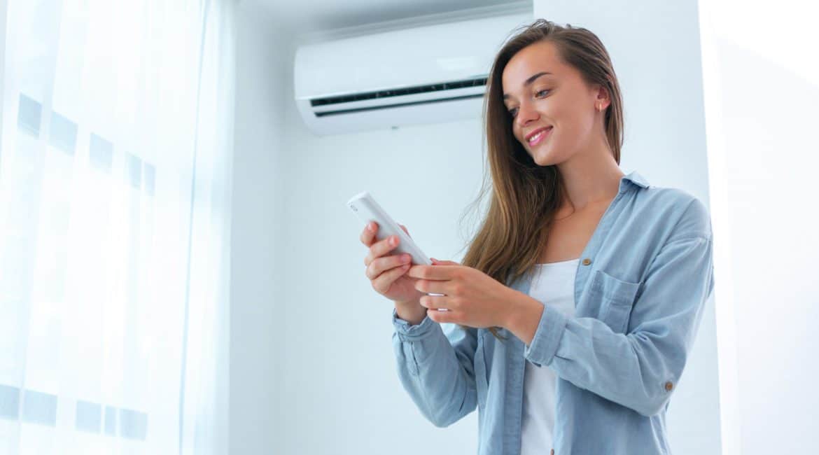 A young woman with light-toned skin and long brown hair adjusts the temperature on the remote for her mini split air conditioner