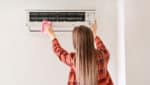A young woman with long light brown hair wiping off the AC unit on her wall with a pink cloth