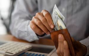 A closeup of a person's hands putting 100 dollar bills into a brown leather wallet