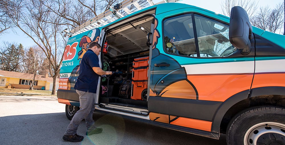 A male Bears Home Solutions heating and cooling technician standing next to his service vehicle in the driveway of a home while looking down at a tablet. 