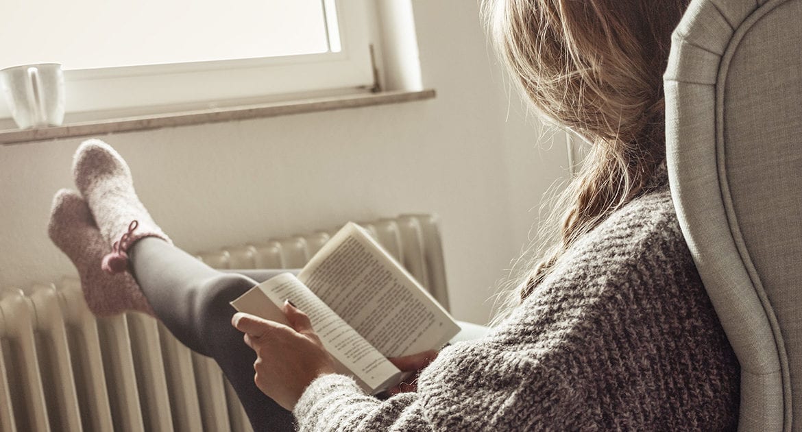 Woman facing the window reads book in chair with feet resting on heat radiator.
