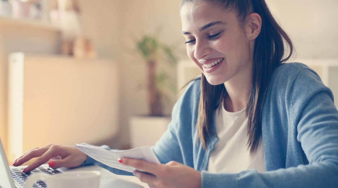 A young woman smiling down at a piece of paper in her left hand while she types on a laptop with her left hand